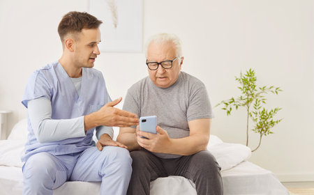 Caregiver helps an elderly man, a patient at a nursing home, use phone. The interaction involves...