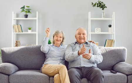 Portrait of smiling gray-haired old couple watching TV together sitting on sofa in living room at...