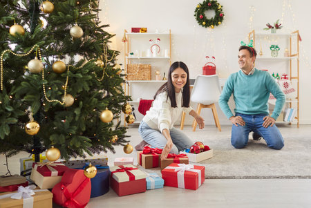 Young happy couple sitting on floor under Christmas tree with many present gift boxes. Cheerful man and woman celebrating New Year together in living room at home. Winter holidays concept.の写真素材
