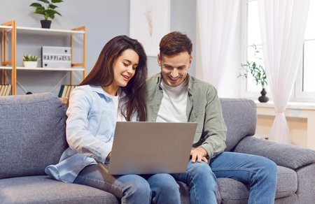 Young family, pleasant couple sitting on sofa in modern living room, looking at laptop screen. Happy pair, married spouse web surfing, making purchases online or booking flight tickets, watching videoの写真素材