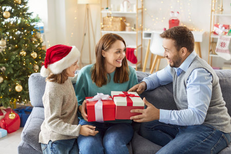 Happy smiling young family congratulating mother on Christmas holiday with present gift box sitting on sofa at home in festive living room. New Year celebration and winter holidays concept.の写真素材