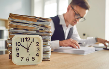 Overworked tired business man accountant working in office sitting at the desk with a clock and a...