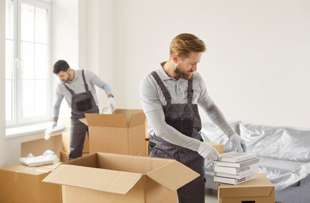 Movers male team packing, choosing cardboard box, arranging items carefully inside, preparing for moving homes or offices, storing, shipping goods and busy organizing items, wrapping to protectの写真素材