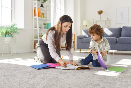 Children and education concept. Mother teaching her little son how to write, preparing for going to school, posing on floor in living room, looking at notebooks, engaged in learningの写真素材