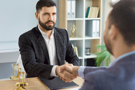 Serious male lawyer shaking hands with man client as sign of agreement, posing at desk in cabinet with scales, discussing legislation issues, getting legal consultation. Legal servicesの写真素材