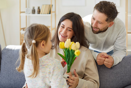Cute small daughter, child girl and loving smilimg young mother, father congratulating with happy birthday or International Womens Day, giving spring flowers bouquet, mothers day gifts at homeの写真素材