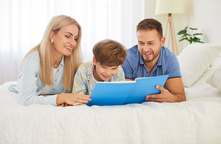 Happy loving family spending weekend home, young father, mother, boy holding reading book, parents with cute little child son, smiling mum, dad telling small kid funny fairy tale story on bed at homeの写真素材