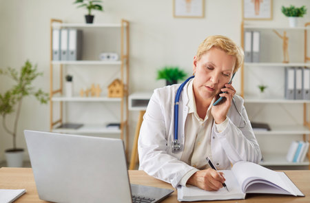 Concentrated elderly doctor or nurse speaking on the phone during call while working online with a laptop in a hospital office. Healthcare, internet connectivity, and professional dedication.の写真素材