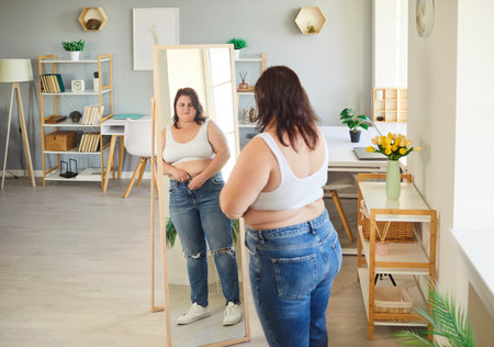 Plus size fat overweight young woman wearing casual clothes looking in the mirror in living room at home. Portrait of plump girl standing indoors. Slimming, diet and obesity concept.の写真素材