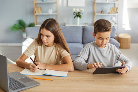 Brother and sister studying together at home, busy doing homework during distance education, using modern technologies to surf necessary information online, posing at desk in living roomの写真素材