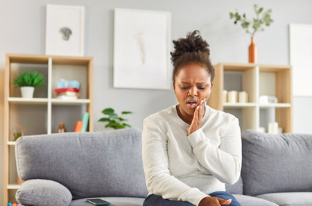 Young African American woman with caries sitting on sofa on living room background at home, holding her hand on her cheek, suffering from discomfort and strong intense tooth pain. Toothache conceptの写真素材