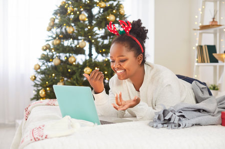 Cheerful young African American woman lying on bed with laptop, making online video call during Christmas Eve. Positive black girl celebrating x-mas at home, congratulating friends or relatives onlineの写真素材