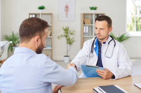 Confident young male doctor therapist in white uniform shaking hands with a man patient sitting at the desk in office after medical examination in clinic. Medicine and health care concept.の写真素材