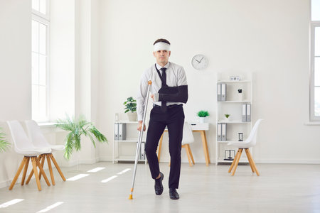 Portrait of a young injured man standing in office with his arm in a plaster cast and bandaged head after accident. Patient wearing sling or bandage walking with crutch. Recovery and rehab concept.の写真素材