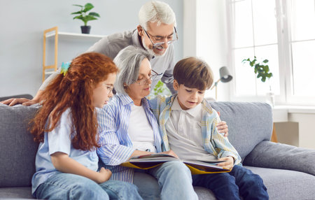 Grandmother reading a book to her grandchildren on the couch at home, close-up. Grandfather and grandmother teach grandchildren to read. Concept enjoy together, good relations, grandparentsの写真素材