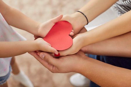 Cropped shot of loving family stacking hands together and holding red paper heart. Close up of mother, father and little child standing close, promoting unity, love, support and strong family bonds.の写真素材