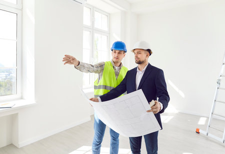 Two men in helmets with a drawing of the building inspect the empty room. The foreman shows the work done according to the plan. The contractor checks the work. Concept constructionの写真素材