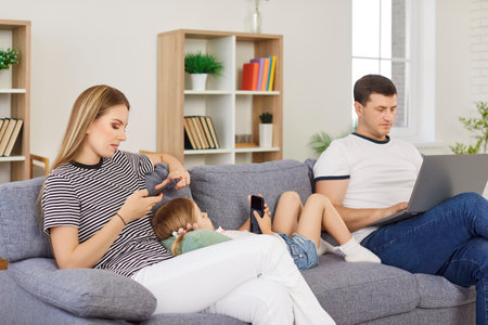 Young family spending time on sofa together, using laptop and smartphones. Parents sitting on sofa with their daughter lying between them, surfing internet, relaxing and enjoying leisure time at home.の写真素材