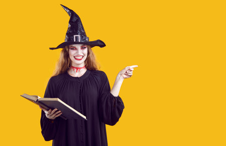 Woman in witch costume pointing finger at copy space, recommending something on Halloween theme. Smiling woman in witch hat, black dress, make-up and spell book on orange background. Web banner.の写真素材