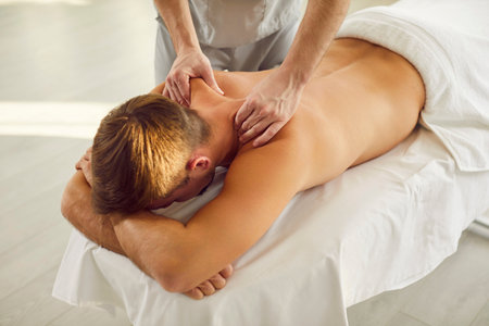 Young man client lying on massage table receiving remedial body back and shoulder massage from professional masseur or doctor physiotherapist in clinic. Physiotherapy and rehabilitation concept.の写真素材