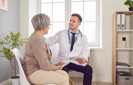 Senior patient in medical clinic talking with male young friendly physician wearing stethoscope and holding report file with appointment during medical examination. Doctor support concept.の写真素材