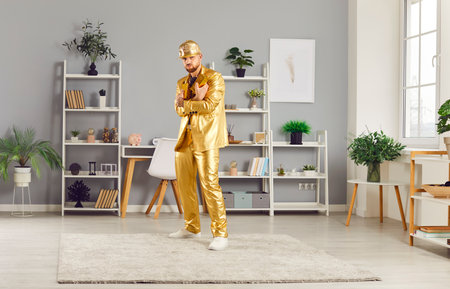 Full length portrait of confident funny young man holding gold bar standing in living room at home. Rich guy wearing golden suit and cap looking cheerful at camera indoors. Business success concept.の写真素材