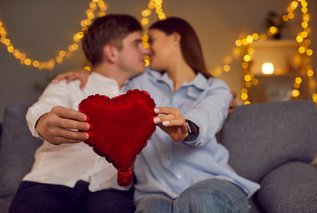 Young cheerful smiling brunette woman and man holding balloon heart in hands looking at each other sitting on sofa at home. Happy couple in love. Romantic, relationship and Valentines day concept.の写真素材