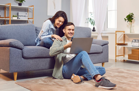 Young laughing couple sitting on sofa in living room at home together watching funny video or using laptop for internet and social media. Happy man and woman resting on couch enjoying weekendの写真素材