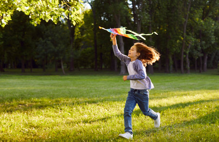 Joyful kid running across green field and holding colorful kite high in air. Happy energetic preteen child enjoying carefree summer in nature, playing with wind during warm sunny day outdoors.の写真素材