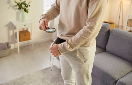 Curious faceless man looking under pants with magnifying glass, has some problems with mens health, posing against home interiorの写真素材