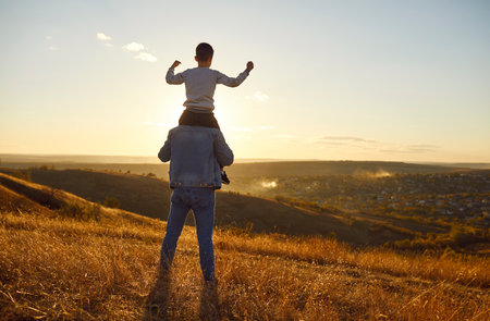 Back view of a little child boy sitting on his fathers shoulders looking into the distance enjoying sunset and beautiful nature outdoors. Son walking with dad in the field. Fathers day conceptの写真素材