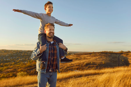 Little happy child boy sitting on his fathers shoulders looking into the distance enjoying nature with copy space. Smiling young father walking with his son in the field outdoors. Fathers day conceptの写真素材