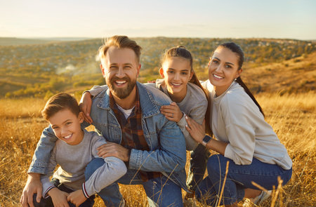 Family with children snuggled tightly together enjoying carefree moment together in golden hour. Portrait of father, mother, son and daughter smiling against background of beautiful natural landscape.の写真素材