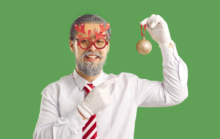 Handsome man dressed in festive clothing, pretending being Santa Claus, pointing at Christmas tree decoration, isolated over green background. New Year is coming soon, think about decorの写真素材