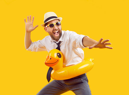 Happy corporate office employee having fun on summer vacation. Joyful, funny, excited, bearded man in shirt, tie, holiday sunhat, yellow rubber duck ring and sunglasses playing on beach. Studio shotの写真素材