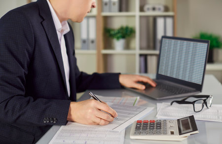 Portrait of a confident young attractive business man accountant in formal suit sitting at the desk looking seriously at laptop monitor screen with charts working in the office on his workplace.の写真素材