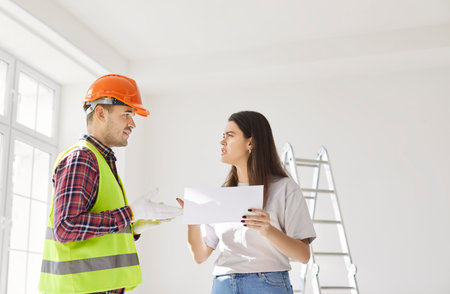 Foreman worker and serious female client asking, directs worker on meeting looking at project plan, holding blueprints, planning work, discussion with construction engineer in hardhat, architect talkの写真素材