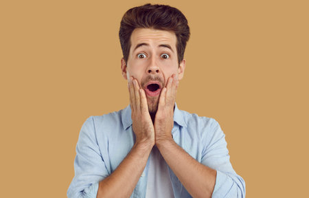 Portrait of young amazed scared man in blue shirt on beige background looking at camera. He touching...