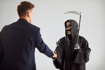 Contract with death. Serious creepy Grim Reaper and young man in suit shake hands after making deal. Skeleton in mantle and with scythe shakes hands with businessman against background of white wall.の写真素材