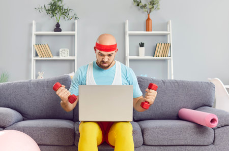 Funny young sportsman sitting on couch, holding fitness dumbbells and looking at monitor. Man with serious facial expression doing exercises, distant training watching a workout online on his laptop.の写真素材