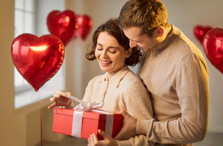 Valentine day gift box giving, love feeling, romantic surprise, happy loving couple in tender hug, kiss, man, woman, heart shaped red balloon foil decor, wedding party room decoration, making proposalの写真素材