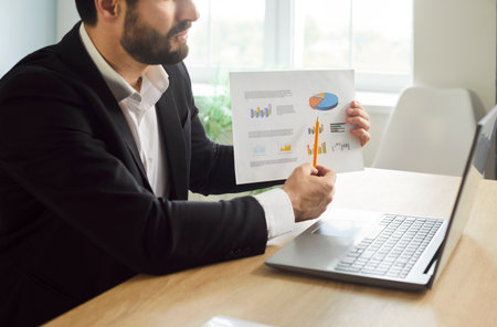 A young businessman working in office showing finance stat at laptop monitor screen having video call sitting at the desk on workplace. Confident man chatting online or having video conference.の写真素材