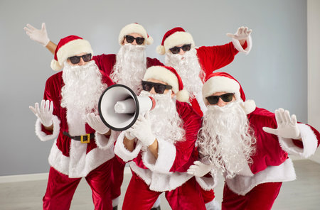 Portrait of a group of excited funny people wearing Santa Claus red costumes shouting announcement in megaphone standing on gray background. Christmas, New year sale and advertising concept.の写真素材