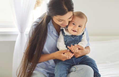 Portrait of happy cheerful smiling young mother hugging her cute little baby sitting on bed at home. Joyful woman with child indoors. Mothers day, maternity, love and care concept.の写真素材