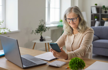 Senior woman pensioner sitting at desk working with laptop at home looking at mobile phone trough glasses having farsightedness eye disease. Mature female person using smartphone on workplace.の写真素材
