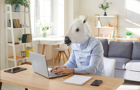 Funny business man and company employee wearing shirt and animal horse mask sitting at the desk on his workplace and working on a laptop in office. Entertainment, fun and fools day concept.の写真素材