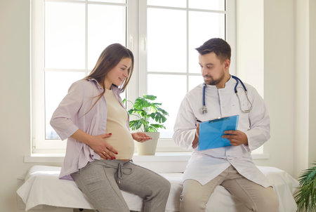 Pregnant young woman sitting on hospital bed and talking with male doctor during scheduled prenatal checkup. Medical specialist listening and taking notes during consultation in medical office.の写真素材