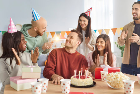 Smiling young happy diverse people group sitting at festive table with cake in the living room...