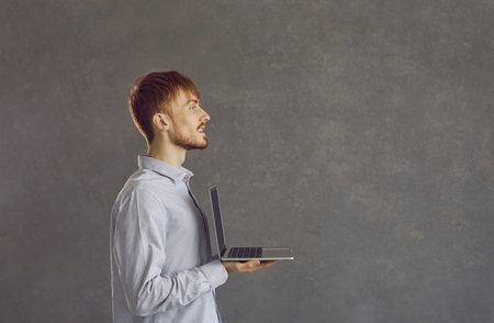 Smiling millennial gen z Caucasian guy isolated on grey studio background hold modern laptop gadget....