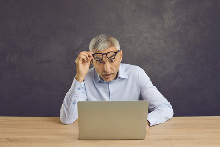 Portrait of shocked senior man who raises his glasses in surprise looking at laptop screen. Man sitting at table on gray background surprised by content or news on Internet.の写真素材